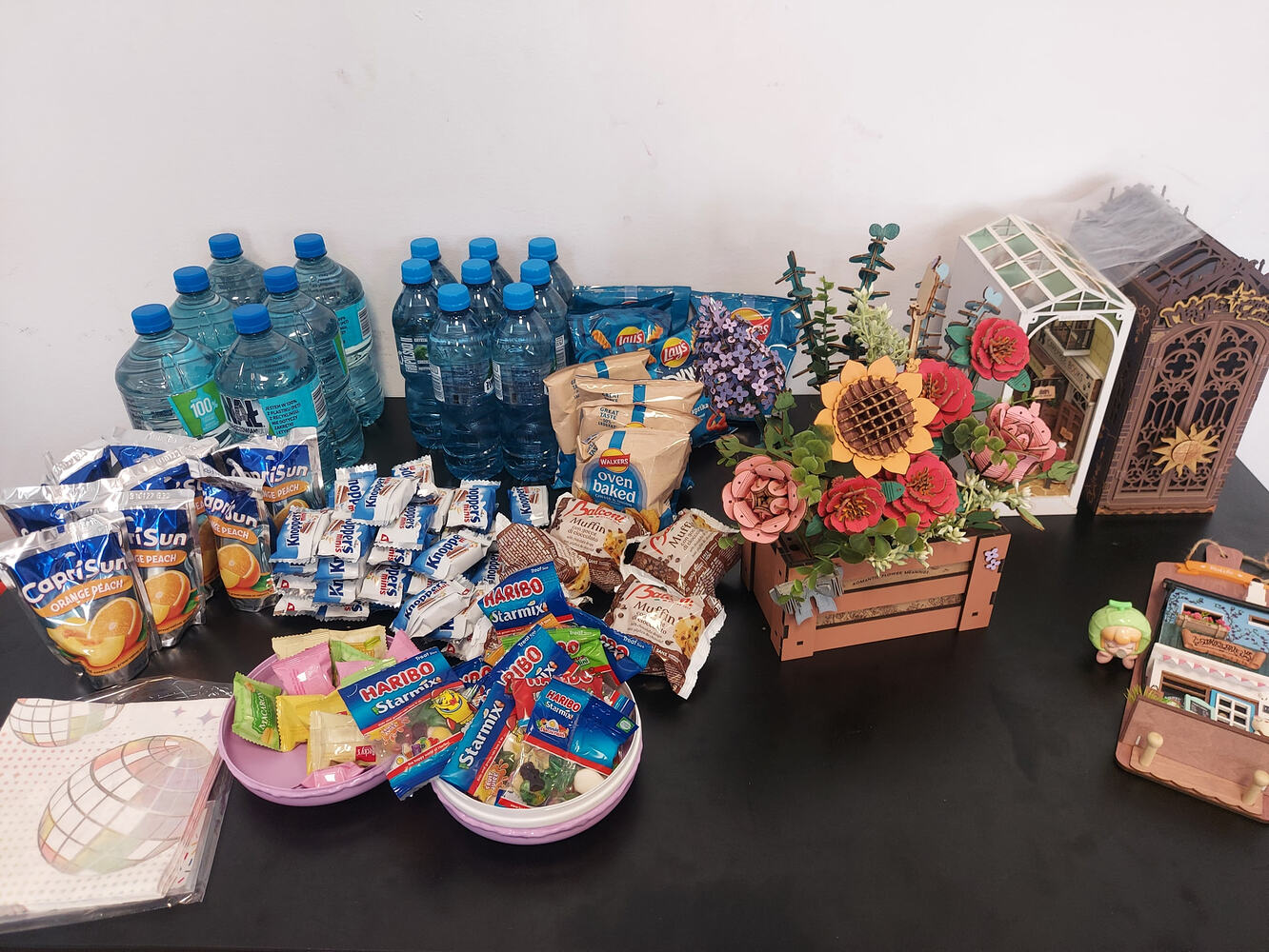 Drinks and snacks arranged beside finished wooden flowers, a greenhouse, a book nook, and a miniature shop